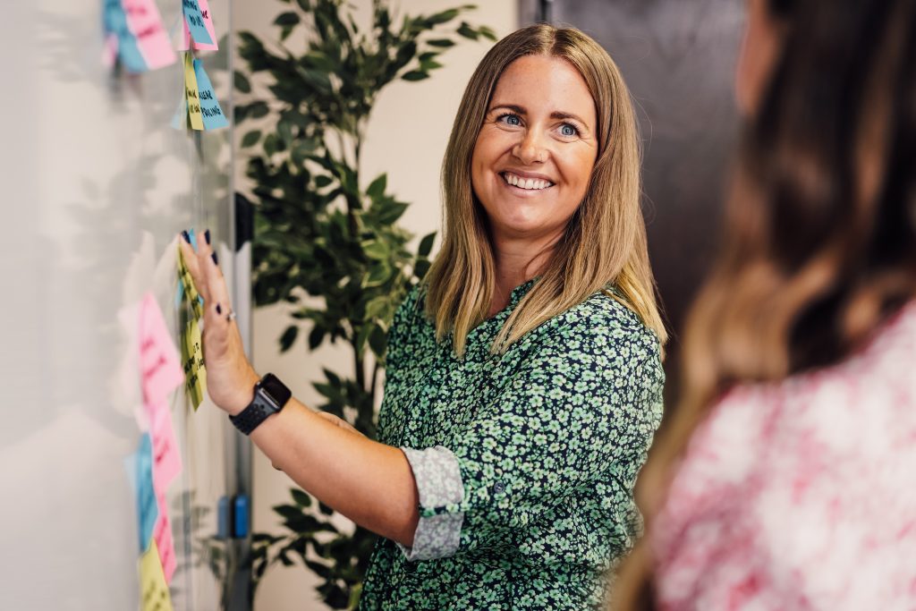 Claire, Director of TMLA, sticking post it notes on a whiteboard as part of a brainstorm, smiling at her colleague.