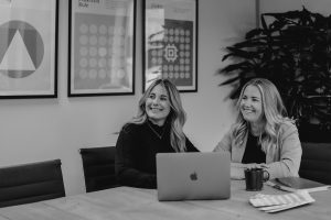 Anna and Claire, Directors of TMLA, sat at a table with a laptop infront of them. They are both smiling.