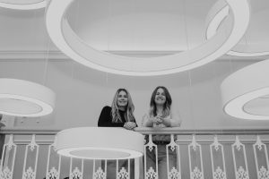 Anna Hutton (left) and Claire MacDonal (right), Directors of TMLA, stood next to each other looking at the camera from a mezzanine floor, with bold circular lighting surrounding them.