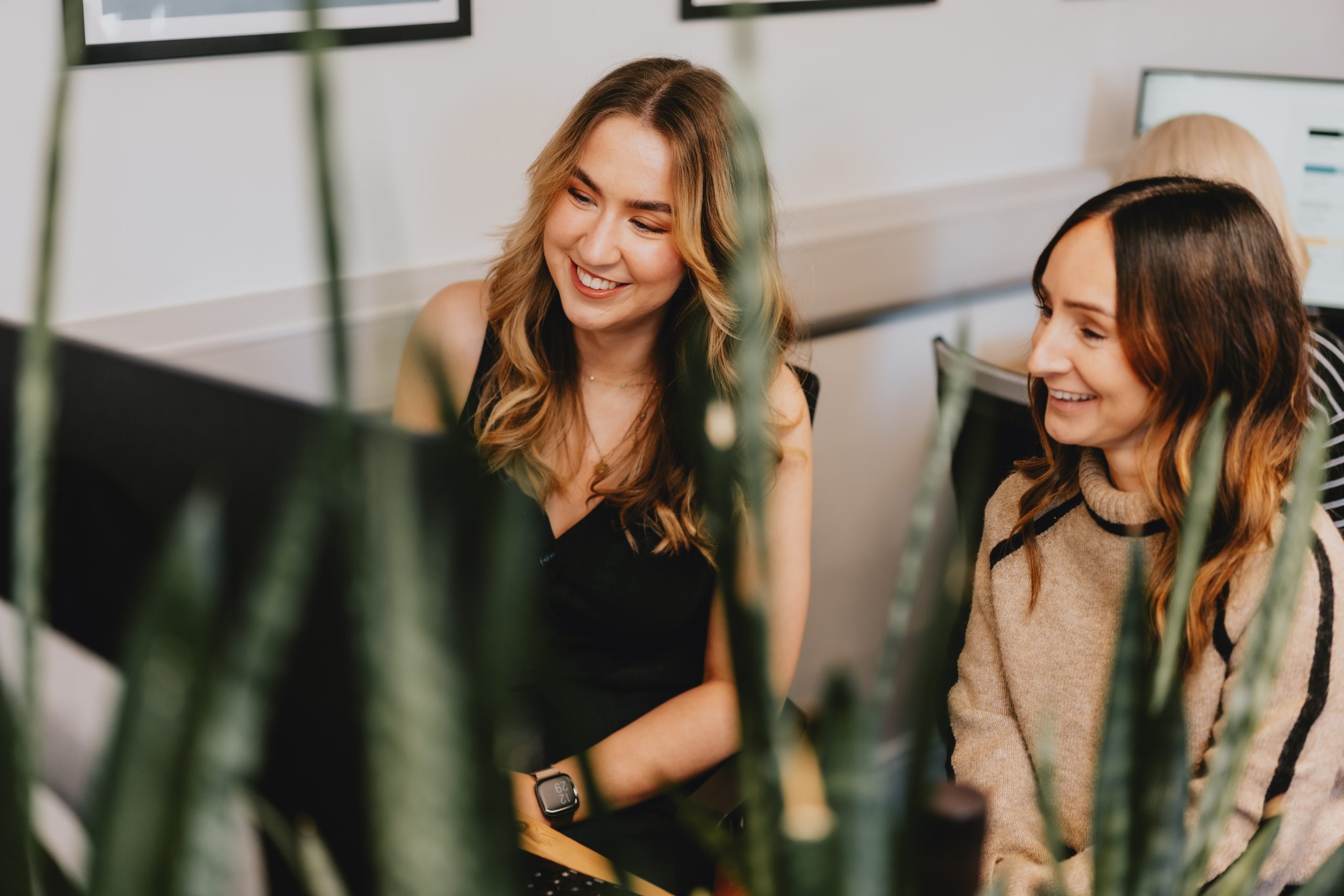 Two female colleagues sat at one desk looking and smiling at a monitor with green plants blurred in the foreground.