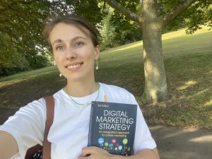 A selfie of Liza, a TMLA student, outside on a walk smiling and holding a book reading 'Digital Marketing Strategy'.